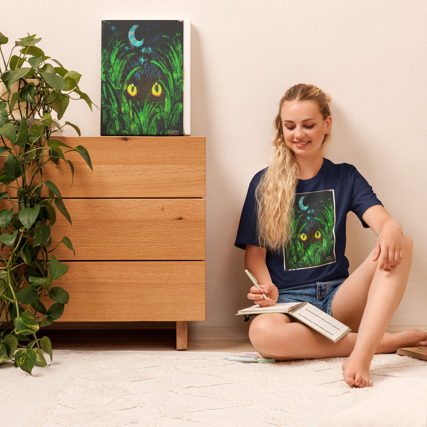 Model wearing navy unisex t-shirt featuring "Midnight Watcher" artwork – glowing yellow eyes in green grass under a crescent moon (Canva Collection)