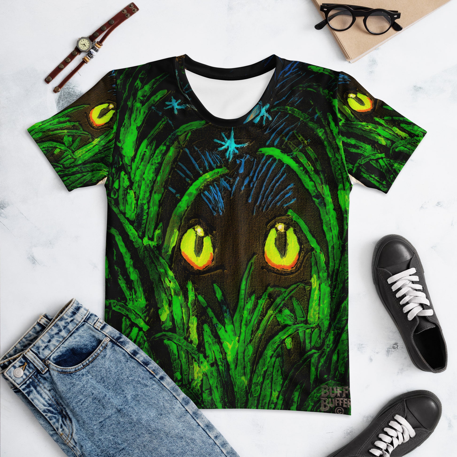 T-shirt with a green and black design of a tree with eyes, surrounded by jeans, shoes, and glasses on a white marble surface.